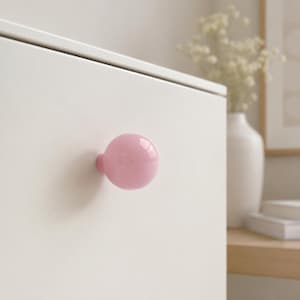May include: A smooth, spherical light pink cabinet knob attached to a flat, matte white surface. The knob has a glossy, reflective finish and a simple, rounded shape that protrudes from the front of the white panel, creating a clean contrast.