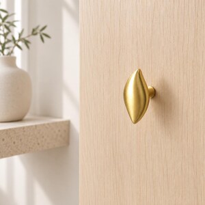 May include: A gold-colored metal cabinet knob with a smooth, elongated, leaf-like shape. The knob features a brushed metallic finish and is mounted onto a light-colored wooden surface, providing a functional handle for furniture or cabinetry.