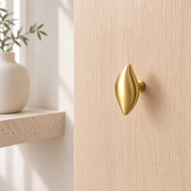 May include: A gold-colored metal cabinet knob with a smooth, elongated, leaf-like shape. The knob features a brushed metallic finish and is mounted onto a light-colored wooden surface, providing a functional handle for furniture or cabinetry.