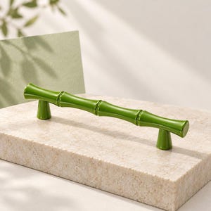 May include: A glossy, grass-green cabinet handle shaped like a segmented bamboo stalk. The hardware features a horizontal bar with three distinct nodes and two cylindrical mounting posts that extend downward from the ends of the handle.