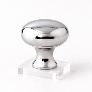 May include: A cabinet knob featuring a polished chrome, mushroom-shaped top attached to a square, clear acrylic base. The metallic surface is highly reflective, showing smooth curves and a bright, silver-toned finish throughout the hardware piece.