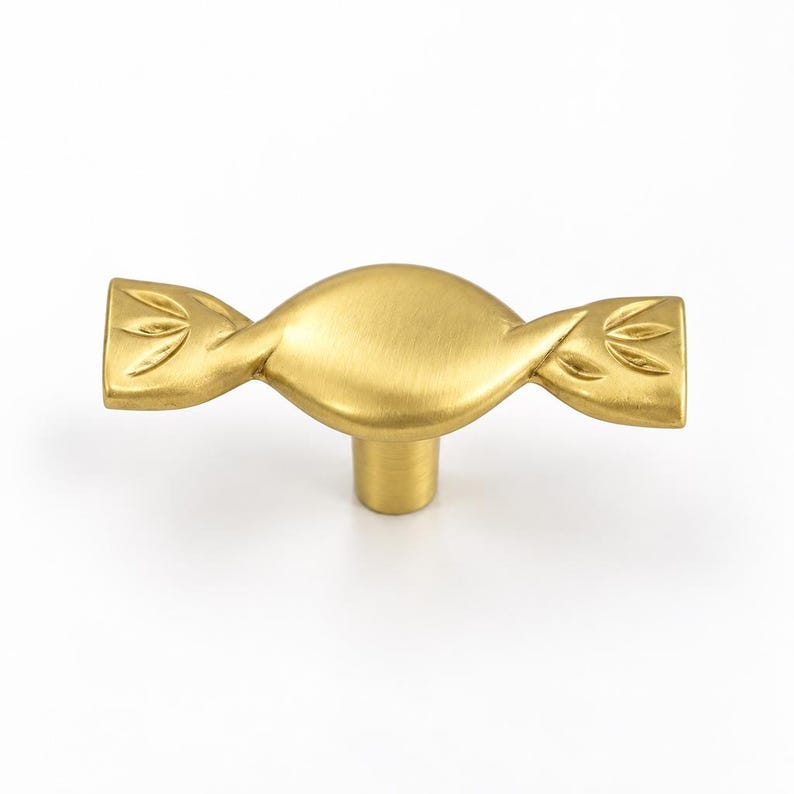 May include: A gold-colored metal cabinet knob shaped like a wrapped candy. The central body is oval and smooth, while the two ends feature a flared, fan-like pattern with etched lines. The hardware has a matte finish and a single cylindrical base.