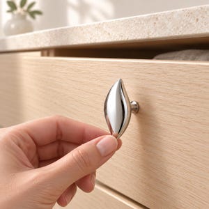 May include: A polished chrome cabinet knob with a smooth, elongated leaf-like shape. The hardware features a reflective, metallic finish and is mounted on a light-toned wood drawer front, showcasing a curved, teardrop-inspired design element.
