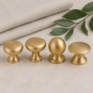 Könnte beinhalten: Vier Schrankknöpfe mit gebürstetem Gold-Finish. Drei der Knöpfe haben eine flache, kreisförmige Oberseite mit einem konischen Sockel, während der dritte Knopf von links eine kugelförmige Oberseite hat. Jedes Stück hat eine glatte Metalloberfläche.