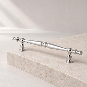 May include: A polished silver-toned metal cabinet pull handle featuring a slender, tapered central bar with decorative spherical and ring-shaped accents at the two mounting points and the ends, creating a symmetrical and elongated hardware piece.