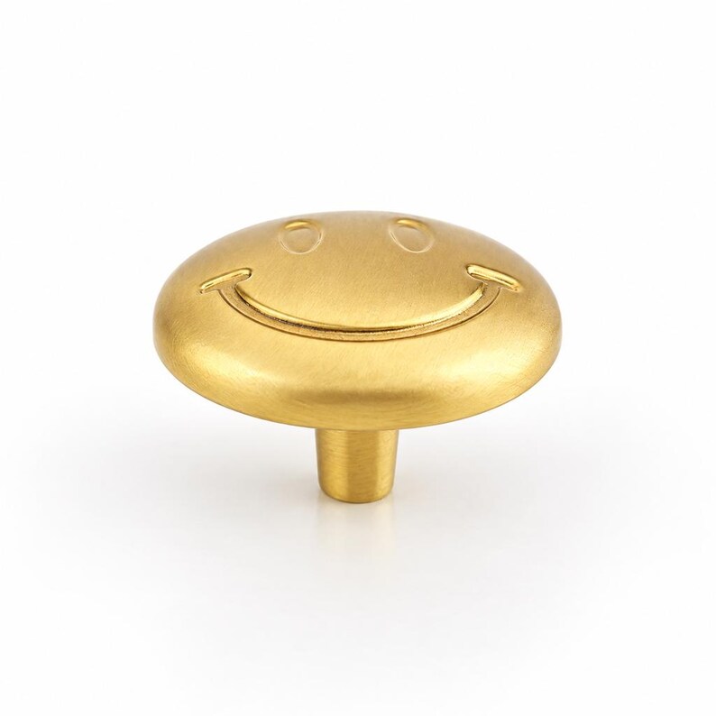 May include: A round, brushed gold metal cabinet knob featuring a debossed smiley face design. The knob has a smooth, circular top surface with two oval eyes and a curved mouth, supported by a cylindrical base extending from the center underside.