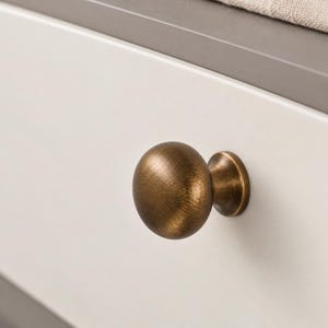 Puede incluir: Un pomo de mueble redondo con acabado en latón antiguo cepillado, que presenta una cabeza esférica lisa y una base cónica, montado en un frente de cajón de color blanco roto con un borde superior gris y un cojín de tela beige al fondo.