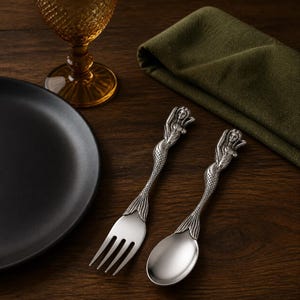 Puede incluir: Un juego de tenedor y cuchara de metal plateado con mangos esculpidos en forma de sirena, con escamas detalladas y una aleta en la base. El tenedor tiene cuatro dientes y la cuchara presenta un cuenco liso y redondeado.