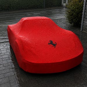 Könnte beinhalten: Eine leuchtend rote Autoabdeckung über einem Fahrzeug mit einem schwarzen springenden Pferd als Logo auf der Motorhaube. Der Stoff ist mit kleinen Wassertropfen bedeckt, was auf nasses Wetter hindeutet, und passt sich der flachen Fahrzeugform an.