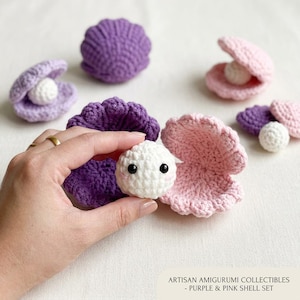 May include: A collection of handmade amigurumi shell sets in purple and pink. Each set includes a crocheted shell and a white pearl with black eyes. The shells are open and closed, creating a whimsical ocean theme.