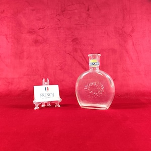 Puede incluir: Una botella de vidrio transparente con cuerpo redondeado y cuello estrecho que presenta una etiqueta Ricard. El vidrio tiene un diseño de rayos de sol en relieve en la parte frontal. Junto a la botella hay una tarjeta blanca sobre un soporte con el texto The French Society.