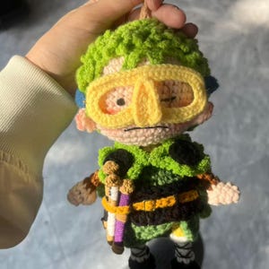 May include: A handmade crochet doll featuring a character with green hair, yellow goggles, and a green and black outfit. The doll has a detailed design with small accessories and is held by a person.