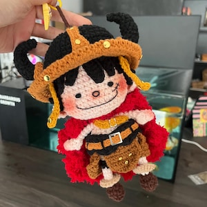 Puede incluir: Una muñeca de ganchillo hecha a mano que representa un personaje con pelo negro, capa roja y falda marrón con cinturón negro. La figura lleva un casco con cuernos negro y tostado adornado con tachuelas doradas y un clip de plástico amarillo.