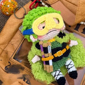 May include: A crochet keychain featuring a character with green hair, yellow goggles, and a green cape. The figure wears a black belt with a gold buckle and carries three cylindrical items in purple, white, and tan. It has white and black leg wraps.