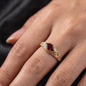 May include: A gold ring featuring a marquise-cut deep red gemstone set in a bezel. The band is crafted with intricate scrollwork and beaded detailing along the edges, providing a textured finish to the metal surrounding the central stone.