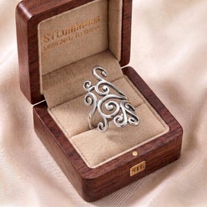 May include: A silver-toned ring with an intricate openwork swirl design resting inside a dark wood box. The interior of the box features a beige velvet lining and the gold-coloured text STLuminous DESIGNED TO SHINE printed on the lid.