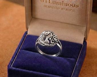 May include: A silver-toned ring featuring a detailed koi fish and lotus flower design, presented inside a tan-coloured box with the text STLuminous DESIGNED TO SHINE printed on the inner lid, resting on a deep blue velvet ring insert.