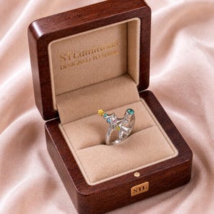 May include: A silver-toned ring shaped like a Christmas tree, featuring small colorful embedded stones and a gold-toned star at the top. The ring is displayed inside a dark brown wooden box with a beige velvet interior and the text STLuminous DESIGNED TO SHINE.