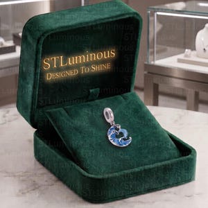 May include: A silver charm featuring a blue and white wave design, displayed inside an open dark green velvet jewelry box. The interior lid of the box features the gold text STLuminous Designed To Shine, with a soft glow emanating from the text.
