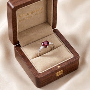 May include: A silver ring featuring a heart-shaped red gemstone flanked by two baguette-cut clear stones, presented in a dark wood box with beige velvet lining. The box lid interior is stamped with the text STLuminous DESIGNED TO SHINE.