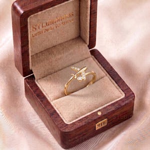 May include: A gold ring featuring an airplane design with small embedded stones, presented inside a dark wood jewelry box. The interior of the box lid is embossed with the text STLuminous DESIGNED TO SHINE, and the front features a small metal tag.