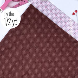 May include: A dark brown fabric featuring a repeating pattern of tiny white dots. The fabric is displayed with a clear plastic ruler and a pink measuring tape, with text on the image reading by the 1/2 yd. Two small red heart pins are visible.