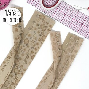 May include: Two strips of beige hook and eye tape featuring a brown floral pattern. The tape includes metal hooks and eyes for garment fastening. A white circular label with the black text 1/4 Yard Increments is visible near the top left.