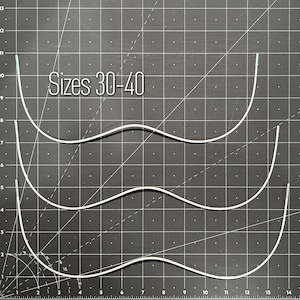 May include: Three white metal underwire bra components arranged vertically. The text "Sizes 30-40" is printed in white above the top wire. The wires are curved with a central dip, positioned against a black grid cutting mat.
