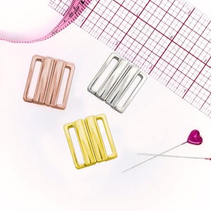 Op de afbeelding: Drie rechthoekige metalen gespen in roségoud, zilver en goud. Een roze meetlint toont aan dat de breedte van de gespen ongeveer 2,5 cm is. Twee dunne metalen spelden met kleine roze hartvormige kopjes zijn inbegrepen.
