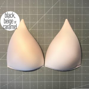 May include: Two beige molded bra cups with a smooth, rounded shape and pointed tops. The text "black, beige or caramel" is printed in a white circle in the upper left corner, indicating available color options for these garment inserts.