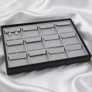 May include: A rectangular black jewellery display tray featuring a light grey velvet interior with twelve individual compartments, each containing small slits for organising earrings. A pair of silver ball earrings is placed in the top left slot.