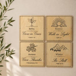 Puede incluir: Cuatro placas cuadradas de madera clara con ilustraciones de líneas negras y texto. Cada placa representa una estación: primavera con un brote, verano con un sol, otoño con hojas de arce e invierno con montañas, incluyendo un versículo bíblico.