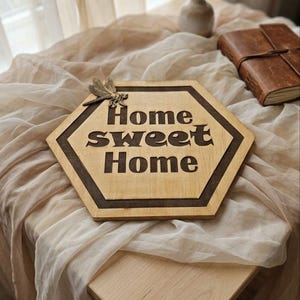 Peut inclure: Panneau hexagonal en bois avec l'inscription « Home sweet Home » gravée en marron foncé. Une libellule décorative est fixée dans le coin supérieur gauche. Le panneau est posé sur une table avec un tissu drapé.
