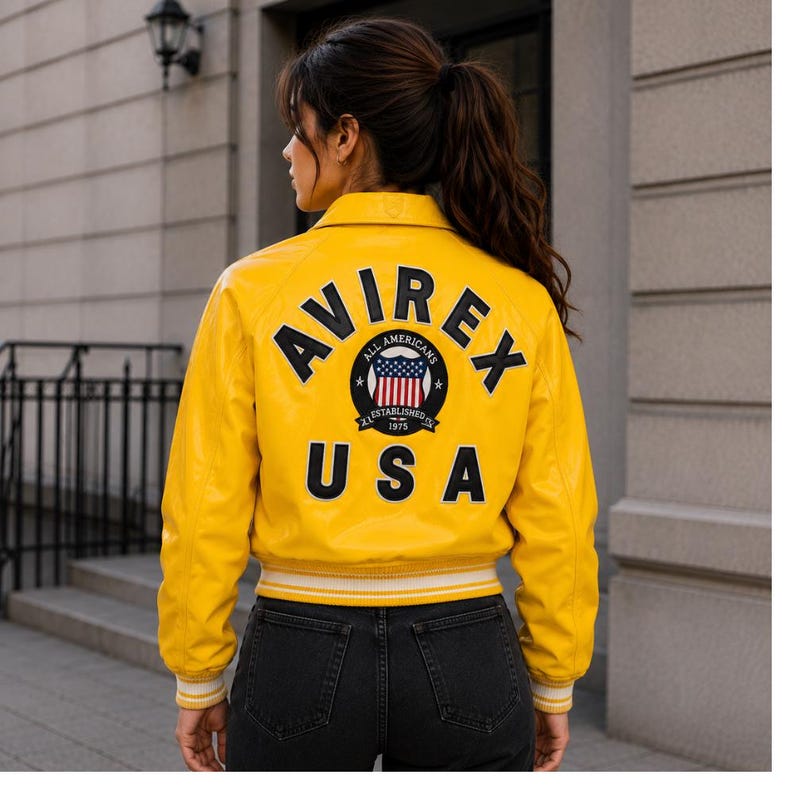 May include: A bright yellow bomber jacket with the text "AVIREX USA" on the back. The jacket features a circular patch with a US flag design and the words "ALL AMERICANS ESTABLISHED 1975". The jacket has a ribbed white and yellow trim.