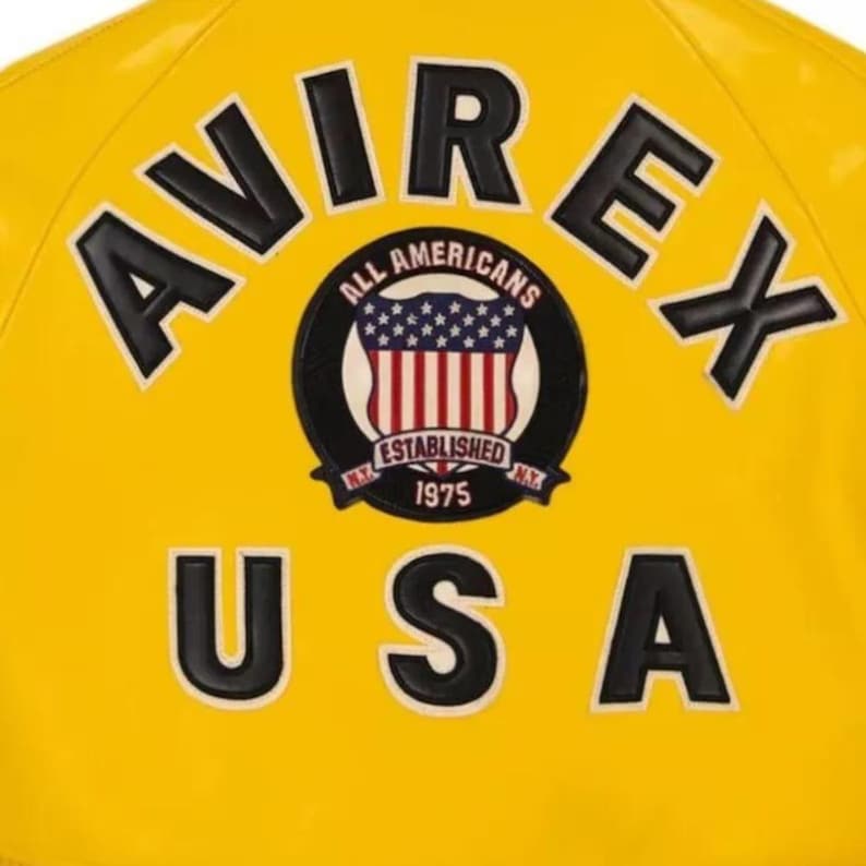 May include: Close-up of a yellow jacket with black lettering and patches. The word "AVIREX" is at the top, and "USA" is at the bottom. A circular patch in the center reads "ALL AMERICANS" and "ESTABLISHED 1975."