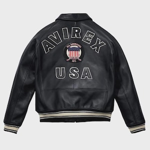 Puede incluir: Chaqueta de cuero negra con la palabra "AVIREX" arqueada sobre los hombros en letras blancas. Debajo, un parche circular con la bandera estadounidense y las palabras "ALL AMERICANS ESTABLISHED 1975". La palabra "USA" est&aacute; debajo.