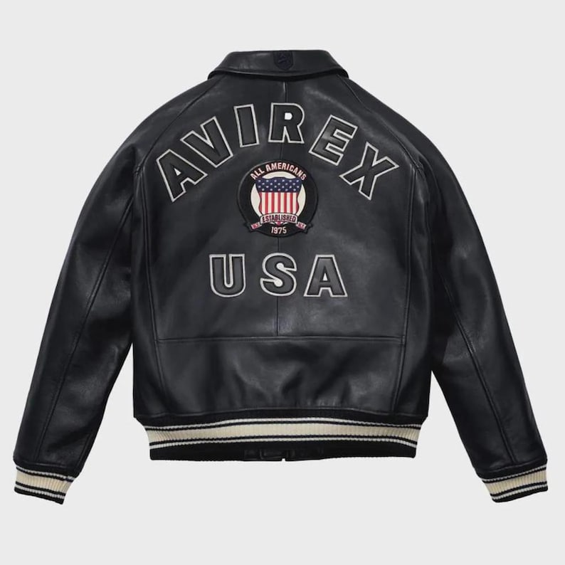 Puede incluir: Chaqueta de cuero negra con la palabra "AVIREX" arqueada sobre los hombros en letras blancas. Debajo, un parche circular con la bandera estadounidense y las palabras "ALL AMERICANS ESTABLISHED 1975". La palabra "USA" est&aacute; debajo.