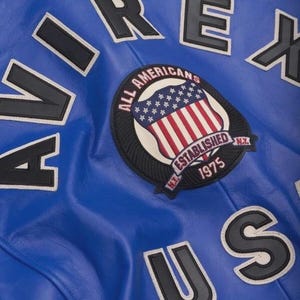 May include: Close-up of a blue leather jacket with white and black lettering. The jacket features a patch with a shield design, the American flag, and the words "ALL AMERICANS ESTABLISHED 1975 N.Y."