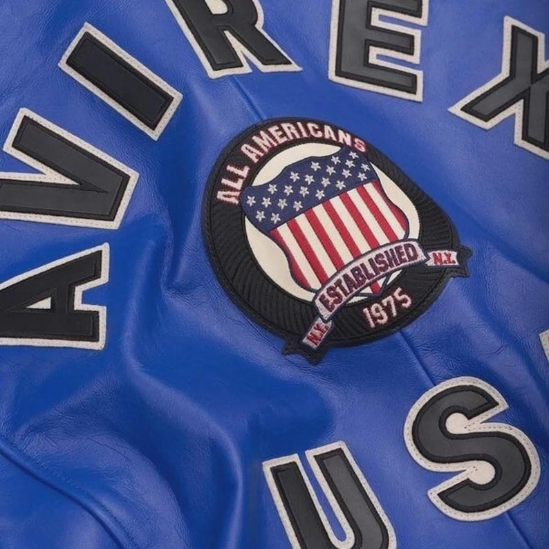 May include: Close-up of a blue leather jacket with white and black lettering. The jacket features a patch with a shield design, the American flag, and the words "ALL AMERICANS ESTABLISHED 1975 N.Y."