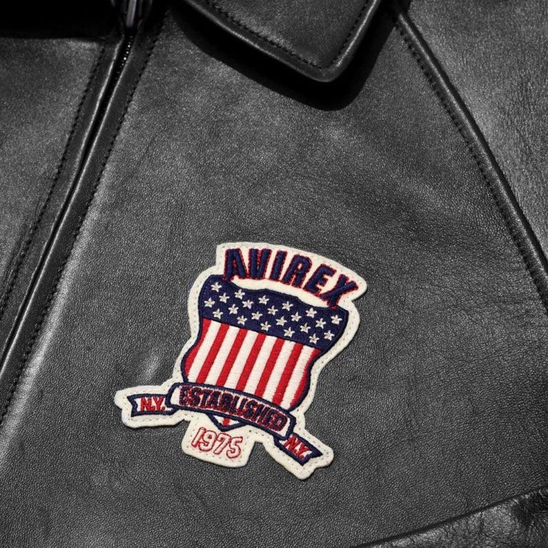 Puede incluir: Primer plano de una chaqueta de cuero negra con un parche. El parche tiene forma de escudo con rayas rojas y blancas, estrellas azules y la palabra "AVIREX". Debajo, el texto dice "NY ESTABLISHED NY 1975".
