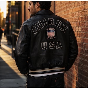 Puede incluir: Chaqueta de cuero negra con texto bordado en blanco y parche en la espalda. El texto dice "AVIREX", "ALL AMERICANS", "USA" y "1975". La chaqueta tiene cuello y pu&ntilde;os acanalados con rayas blancas y beige.