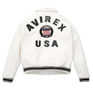 Op de afbeelding: Witte leren jas met zwart-wit gestreepte manchetten en kraag. De achterkant heeft het woord "AVIREX" boven een cirkelvormig embleem met de woorden "ALL AMERICANS ESTABLISHED 1975" en een USA-vlagontwerp. Het woord "USA" staat eronder.