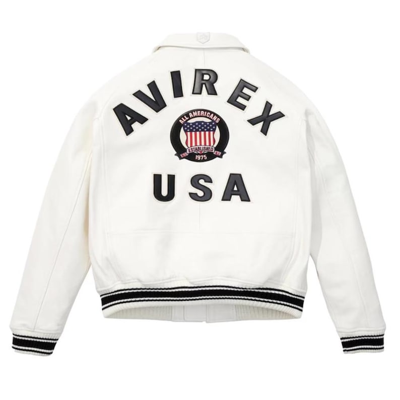 Op de afbeelding: Witte leren jas met zwart-wit gestreepte manchetten en kraag. De achterkant heeft het woord "AVIREX" boven een cirkelvormig embleem met de woorden "ALL AMERICANS ESTABLISHED 1975" en een USA-vlagontwerp. Het woord "USA" staat eronder.