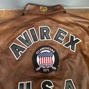 Może przedstawiać: Brązowa sk&oacute;rzana kurtka z napisem "AVIREX" w białych i czarnych literach na plecach. Widoczna jest r&oacute;wnież naszywka z napisem "ALLAMERICANS ESTABLISHED 1975" oraz wzorem amerykańskiej flagi.