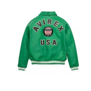 Puede incluir: Chaqueta de cuero verde brillante con la palabra "AVIREX" arqueada sobre los hombros. Debajo, un parche con la bandera americana y la palabra "USA". La chaqueta tiene cuello y ribete a rayas blancas y verdes.