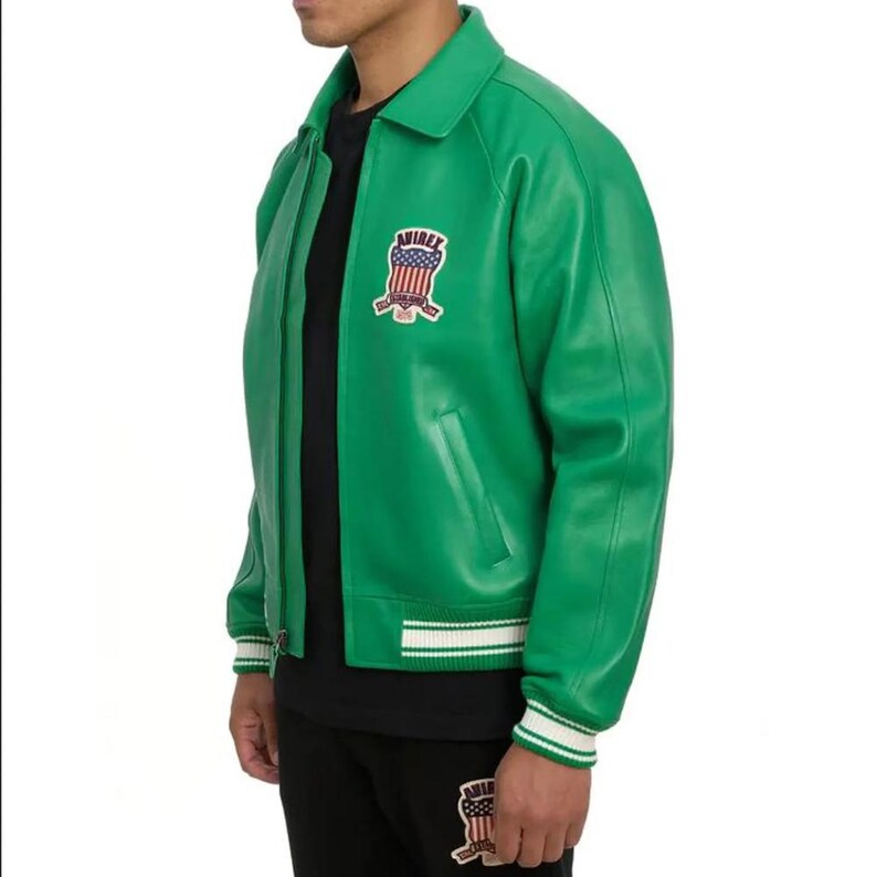 Puede incluir: Chaqueta de cuero verde con cuello, cierre de cremallera y pu&ntilde;os y dobladillo a rayas blancas y verdes. La chaqueta presenta un parche con el texto "AVIREX" y un dise&ntilde;o patri&oacute;tico.