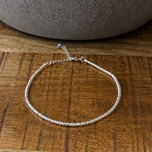 May include: A delicate silver bracelet with a textured design. The bracelet is on a wooden surface, with a grey, rounded object in the background. The bracelet has a small chain for adjusting the size.