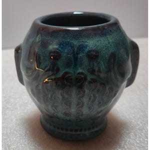 May include: A small, rounded ceramic vessel featuring a deep teal and dark blue glaze with a glossy finish. The exterior surface displays a textured, embossed pattern of swirling, organic shapes and two small, protruding side handles.