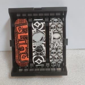 May include: Three finger skateboards displayed in a black plastic rack. The left board is orange with a black plaid pattern and the word BLIND. The centre board is black and white with a cartoon character. The right board features a skull pattern.