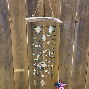May include: A hanging mobile featuring a horizontal piece of driftwood suspended by twine. Multiple vertical strands hang from the wood, adorned with various small white seashells and dark green sea glass pieces. A small star ornament is visible below.